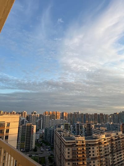 The image shows a view of a densely populated urban area with numerous multi-story apartment buildings. The buildings vary in height and architectural style, with some featuring colorful facades and others presenting more uniform, modern designs. The sky above is partly cloudy with patches of blue visible, suggesting either early morning or late afternoon light. The foreground includes a portion of a balcony railing, indicating the vantage point is from an elevated position. The overall impression is of a large residential district within a city. The location of Feklistovo, Russia, cannot be confirmed from the visual information provided.