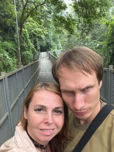 Two individuals, a woman and a man, are captured in a selfie on a raised walkway. The walkway, constructed with metal railings and a slatted surface, appears to be in a lush, green, forested environment. The woman is positioned on the left of the frame, smiling warmly, with brown hair framing her face. She is wearing a light-colored jacket over a dark choker necklace. The man is on the right, his left eye squinting, with light brown hair. He is wearing a tan-colored shirt with a black strap crossing his chest. The background is dominated by dense foliage, with trees and vegetation visible on either side of the walkway, suggesting a natural setting. The location is identified as a walkway in the Mae Sa Waterfall National Park, near Pong Yaeng, Thailand.