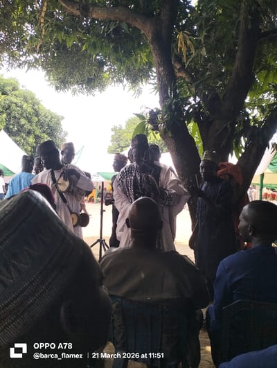 This outdoor scene in Gudi, Nigeria, depicts a gathering on 21 March 2026 at 11:51. A central group of individuals stands under a large, leafy tree. A prominent male figure in a white tunic with dark patterns and a dark cap stands with his hands clasped, appearing to address others. To his left, a man in a white tunic and cap plays two hourglass drums, one held and one suspended. To the right of the central figure, a man in a dark blue garment and cap gestures with an open hand. Additional men in traditional attire and caps are visible standing in the background, some near white canopy tents with green trim. In the immediate foreground, two men are seated with their backs to the camera on blue plastic chairs; one wears a dark garment and cap, the other a dark blue garment. The ground is bare earth, and the scene is brightly lit by daylight, partially filtered by the tree's foliage. A black tripod stand is visible in the mid-left background.