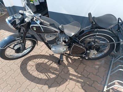A vintage black DKW motorcycle is parked on a brick-paved surface next to a white and dark gray building in Germersheim, Germany. The motorcycle has a single seat with a pillion pad and a luggage rack. The front of the motorcycle features a prominent headlight with a speedometer. The engine casing is visible, bearing the "DKW" insignia. The scene is illuminated by sunlight, casting a shadow of the motorcycle on the ground. A metal grate and a silver bicycle rack are visible in the lower right corner.
