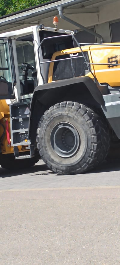 A low-angle, medium shot captures the side of a yellow and white construction vehicle, likely a loader, parked outdoors on an asphalt surface. The vehicle features a prominent, thick tire with a deeply treaded pattern, and a large, black fender arches over it. The cabin of the vehicle is partially visible on the left, with an open door revealing a steering wheel and controls. A small, orange flashing light is mounted on the roof of the cabin. The background includes a building with a gutter and some green foliage. The scene is well-lit by natural daylight, casting a shadow from the vehicle onto the ground. The overall impression is one of an industrial setting.