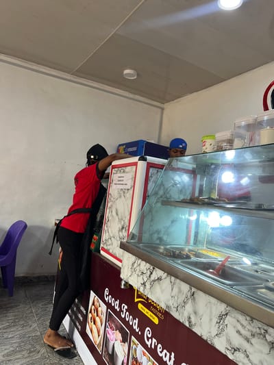 This media file depicts an indoor scene at a food establishment, possibly a bakery or snack shop in Ifite-Awka, Nigeria. The main subjects are two individuals and a display counter filled with food items.
In the foreground, a person wearing a red t-shirt, black apron, and a black cap is partially visible, seen from the waist down. They are standing next to a display counter, and their feet are bare, wearing sandals. The display counter is made of glass and metal and holds various food items, appearing to be snacks or pastries. The counter itself has a marble-patterned base.
Behind the counter, a person wearing a blue cap and a blue t-shirt is seen leaning over the counter, looking at the food items. This individual is the subject of the "snowsea" text which appears to be a label on a blue container.
A prominent feature of the scene is a large banner or menu board attached to the side of the counter. The visible text on this board includes "ATILI VLLIYIII PASTRIES" and "CRAVE, EAT, REPEAT." Further down, it says "Good Food. Creat pastries." The overall impression is that of a small business actively serving customers.
The lighting suggests it is daytime, likely during business hours. The indoor setting appears to be a well-lit commercial space. No specific weather information can be inferred. The overall mood is one of daily business operations within a food service environment.