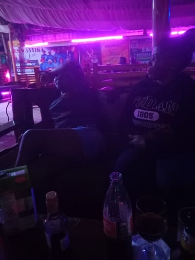 The image shows the interior of a dimly lit bar or nightclub. The scene is dimly lit, with purple neon lights illuminating the room. There is a white fabric canopy or ceiling above, suggesting an outdoor seating area. Two individuals are seated at a table in the foreground. One person is wearing a black shirt with a white logo, while the other is wearing a dark-colored shirt.  The person wearing the black shirt has "VILLAN 1805" printed on their shirt.  There are two bottles on the table. One appears to be glass and the other is an opaque brown plastic bottle. A glass of wine is positioned between the bottles.  A third person is visible behind the first two seated individuals, their face obscured by the shadows.  A sign on the wall reads "ES WANYIKA Live" and  “BUNGOMA RHUMBA.”  The scene is located in the city of Bungoma, Kenya, specifically at the coordinates 0.593 latitude and 34.559 longitude.  The scene suggests a social gathering involving drinks and conversation.  The overall atmosphere is relaxed and convivial.