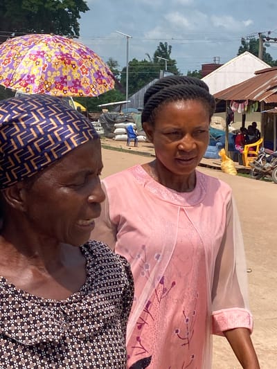 Two women are walking down a dirt road in Igbara, Nigeria. The woman in the foreground on the left is wearing a dark patterned headscarf and a dark patterned blouse. She is looking to her right. The woman on the right is wearing a pink dress with a sheer overlay. She is looking forward and slightly to her right. In the background, there is a colorful umbrella, stacks of white bags, a blue chair, and a motorcycle. Buildings with corrugated metal roofs are visible. The sky is cloudy.
