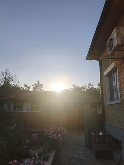 The image depicts a serene outdoor scene, possibly a backyard, in Harmanli, Bulgaria. The sky is a pale blue, with the sun shining brightly, creating a lens flare that obscures some of the details. Two buildings are visible, one with a tiled roof and windows, and another more modern structure with an air conditioning unit mounted on the exterior. The foreground features a paved walkway and a garden with pink flowers and greenery. The overall impression is one of a peaceful residential setting, likely captured during sunrise or sunset.