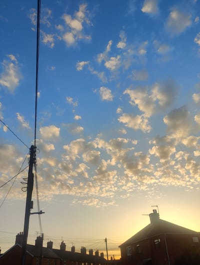 The image captures a suburban scene, likely in East Suffolk, United Kingdom, suggested by the architectural style of the terraced houses. The foreground is dominated by a utility pole with electrical wires stretching across the frame. The lower portion of the image includes rooftops of terraced houses with visible chimneys and TV antennas. A detached house is visible on the right side, with the sun setting behind it, casting a warm glow. The sky is filled with a mix of clear blue and small, puffy white clouds, illuminated by the setting sun. No people are visible. The scene suggests a quiet residential area at dusk.