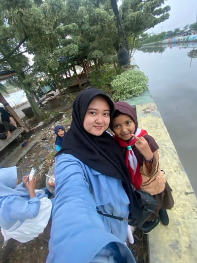 The image captures an outdoor scene in Kalitengah, Indonesia, featuring an adult female and a child posing for a selfie alongside a body of water. The adult female, positioned in the foreground and appearing to hold the camera, has a light complexion, wears a black hijab, and a light blue long-sleeved top; she smiles directly into the lens. To her right, a young girl in a brown head covering (hijab), a brown and tan uniform with a red and white neckerchief, and a black shoulder bag, also smiles at the camera while making a "rock on" hand gesture with her right hand, revealing a purple bracelet.

The background on the right side of the frame shows a wide body of water, possibly a river or canal, reflecting the overcast sky. The water's edge is a yellow-grey concrete or paved barrier. Across the water, a distant wall with a colorful mural is discernible. Near the concrete edge, a bush with small white flowers is visible, and a dark, bell-shaped object hangs from an overhead structure or tree branch.

On the left side of the background, a smaller child in a blue hijab and top looks towards the camera, partially obscured. Further to the left, several individuals are seated on a low, makeshift platform constructed from corrugated metal and wood. One person in a light blue hijab is engaged with a smartphone. The ground beneath this seating area appears unkempt with visible debris. Lush green trees with dense foliage dominate the upper background, partially obscuring a wooden gazebo structure.