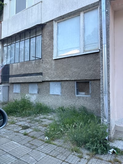 The image depicts the exterior of a multi-story building, likely an apartment block, with a rough, textured concrete facade. On the ground floor, several small, square openings are visible, some covered with what appear to be metal grates or panels. Tall, wild grass and weeds grow in abundance along the base of the building and on the paved walkway in front of it. The paving stones are uneven and cracked, with vegetation pushing through the gaps. To the right, a silver downspout runs vertically along the corner of the building. The overall impression is one of neglect and disrepair, common in older residential areas.
The scene is located in Silistra, Bulgaria, a city on the Danube River known for its historic remnants and Soviet-era architecture. This particular view likely captures a section of a more utilitarian, post-war era housing complex, characterized by its functional design and durable, albeit unadorned, materials. The overgrowth of vegetation suggests a lack of regular maintenance or landscaping. The presence of a car mirror in the lower left corner indicates the photograph was taken from a vehicle, possibly documenting the condition of the street or a specific building.