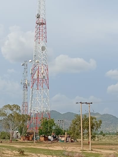 A wide-angle, outdoor, full shot captures a telecommunications tower and a smaller, adjacent tower in Shuwa, Nigeria. The main tower, painted white and red, is the dominant feature, with several circular dishes mounted on its structure. To its left, a second, thinner tower also supports communication equipment. In the foreground, scattered trees and small, rustic huts with thatched roofs are visible. In the background, rolling hills with sparse vegetation form a natural backdrop under a partly cloudy sky. The scene is characterized by a dry, open landscape, suggesting a rural or semi-rural setting within Shuwa.