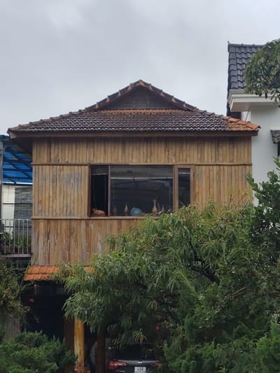 The image shows a two-story wooden house with a tile roof in Lang Biang - Đà Lạt, Vietnam. The house has a large window on the second floor, which is partially obscured by trees. Inside the window, there are several vases and a globe. The first floor of the house appears to be an open-air parking space, with a car visible. The house is surrounded by lush greenery, and the sky is overcast. The overall impression is one of a rural or semi-rural setting.