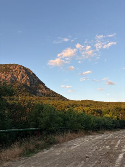 The image depicts a mountainous landscape with a dirt road in the foreground. The sky is blue with scattered clouds, some of which are tinged with pink from the setting sun. A large, rocky mountain dominates the left side of the frame, its slopes covered in dense green foliage. The middle ground is characterized by a rolling hill covered in trees, with the warm light of the setting sun illuminating the upper portions of the foliage. A thin, green pipe runs horizontally across the middle ground, partially obscured by vegetation. In the far right of the image, a small, pale circle, likely the moon, is visible in the sky above the tree-covered hill. The dirt road in the foreground is uneven and appears to be recently disturbed, with tire tracks and scattered stones. The overall impression is of a natural, rural environment at dusk.