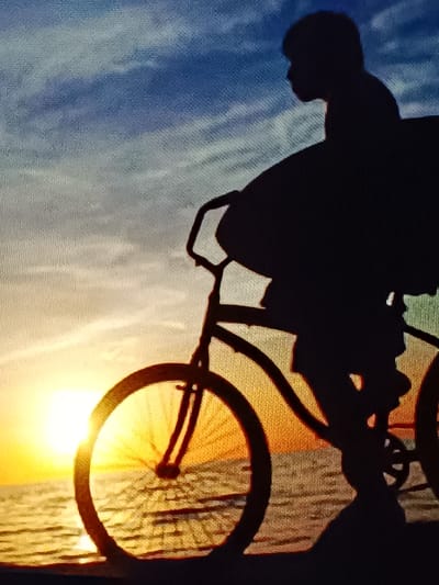 A silhouette of a person riding a bicycle with a surfboard is captured at sunset. The scene is set against a backdrop of the ocean and a colorful sky with shades of blue, orange, and yellow. The sun is low on the horizon, casting a warm glow and reflecting on the water. The bicycle appears to be a cruiser style with large wheels. The surfboard is positioned behind the rider, leaning against the bicycle. The overall impression is one of a peaceful and serene moment at the end of the day. This scene does not depict any location within the city of Vitoria-Gasteiz, Spain.