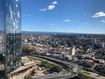 This image captures a panoramic view of a city, dominated by a modern, cylindrical skyscraper on the left. The skyscraper is clad in reflective blue glass, mirroring the bright blue sky and white clouds above. The city spreads out beneath a clear, sunny sky, extending to a range of hazy blue-grey mountains on the horizon.
The urban landscape consists of a dense mix of buildings, ranging from low-rise residential structures with varying rooflines to taller, more modern buildings in the distance. A prominent feature is a wide, elevated road or overpass that curves through the mid-ground, appearing to follow a waterway or dry riverbed flanked by trees and greenery. Along the banks of this waterway, several buildings display colorful facades and visible graffiti.
At the base of the skyscraper, there is an ongoing construction site with dirt, some machinery, and a white van. Another white van is visible on the road in the foreground, and in one of the cropped hints, two people can be seen walking on the elevated road, while another person cycles on an adjacent path.
The time of day is daytime, likely late morning or early afternoon, characterized by bright sunlight casting distinct shadows. The weather is clear and sunny with scattered cumulus clouds. The location is Cordoba, Argentina.
Visible text includes a series of blue signs with white social media icons (Facebook, Twitter, YouTube, Instagram) mounted on a structure at the base of the skyscraper, below which is a white banner with indistinct dark text. Graffiti is also visible on some building walls along the riverbed.