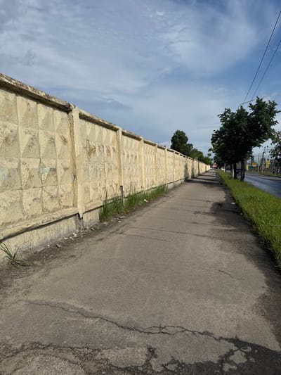 A ground-level, vertical shot showcases a cracked, yellowed concrete wall running diagonally from the lower left corner towards the upper right. The wall has a textured, repetitive diamond pattern. To the right of the wall is a paved sidewalk that appears wet in places and shows signs of disrepair with cracks and patches of darker asphalt. On the other side of the sidewalk, there is a strip of green grass and then a road. Beyond the road, more trees and what appear to be utility poles and overhead wires are visible, suggesting an urban or suburban environment. The sky above is a mix of blue and white clouds, indicating a partly cloudy day. The lighting suggests it is daytime. No people are visible in the frame.