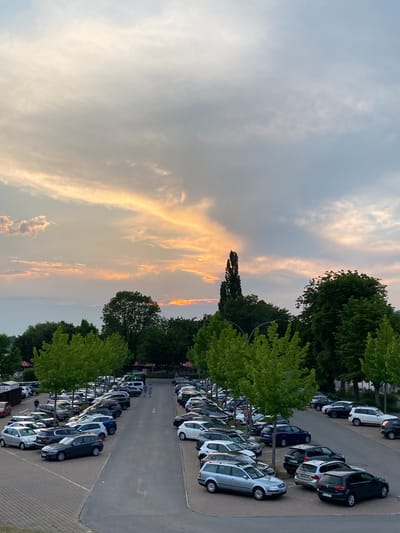 The image shows a full parking lot at dusk in Rinteln, Germany. The sky is filled with dramatic clouds, with orange and yellow hues indicating the setting sun. Rows of parked cars, predominantly sedans and SUVs, occupy the asphalt lot. Several deciduous trees with vibrant green foliage are planted between parking sections and line the edges of the lot. In the background, a cluster of darker green trees forms a treeline. Two individuals are walking away from the camera down the center of the parking lot. The overall scene depicts a typical evening in a town, with the natural beauty of the sky contrasting with the mundane presence of a busy parking area.