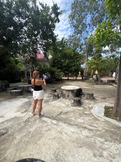 This image captures a bright, sunny daytime scene in what appears to be a temple or garden complex in Cam Ranh, Vietnam. The main subject is a person, seen from behind, standing on a concrete path. They are wearing white shorts, a black sleeveless top, and white Crocs, with their hair in a braid and sunglasses on their head. The person is holding a smartphone up, seemingly taking a photo or video of the area in front of them.
The setting is characterized by a mix of natural elements and man-made structures. The ground is largely concrete, showing signs of wear and some cracks, with occasional patches of soil and grass. To the right of the person, there's a stone or concrete table and two stools, carved to resemble tree stumps.
The background is dominated by a variety of green trees and lush foliage under a clear blue sky dotted with scattered white clouds. Amidst the trees, traditional Asian architecture is visible, including a building with a prominent red-tiled roof and white ornate decorative elements. A swastika symbol, commonly found in Buddhist temples, is discernible on this structure. Further into the mid-ground, partially obscured by greenery, at least two golden Buddha statues can be seen, reinforcing the impression of a religious site. The overall mood is peaceful and serene. No explicit text is visible in the image.