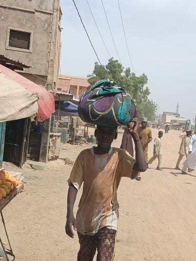 A young male is depicted walking forward on an unpaved, light brown dirt road in Hotoro North, Nigeria. He is positioned centrally, wearing a soiled light-brown and white striped short-sleeved shirt and dark brown trousers with a repeating light-colored pattern. A large, bulbous bundle, wrapped in multicolored fabric with teal, purple, black, and white patterns, is balanced on his head, stabilized by his raised right hand. His gaze is directed towards the viewer with a neutral expression.
To the immediate left, a market stand displays a pile of ripe yellow-orange fruit, likely mangoes, on a wooden surface. Behind this stand, a two-story concrete building, appearing unfinished or aged, shows an exposed square window opening. Below this, a structure with a red and beige tarp awning is visible. Further left are additional basic market stalls with corrugated metal components.
In the mid-ground, several male figures are walking on the same road. One individual in a brown garment and cap is partially visible behind the main subject. Further along, a young boy in a light-colored tunic walks, and on the far right, a man in a white thobe and cap is also in motion.
The background features additional buildings of varying heights and materials, including concrete and corrugated metal, suggesting an urban or semi-urban commercial area. Green trees are visible beyond the structures on the right. Multiple dark power lines extend across the pale, hazy sky, indicative of a bright, possibly dusty, day.