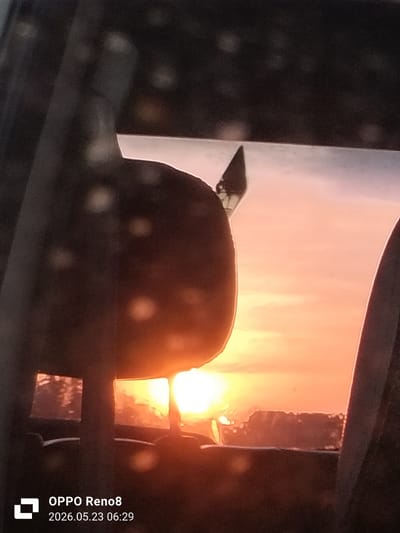The photo, taken on May 23, 2026, at 06:29, presents a blurred view of a sunrise, seemingly from inside a vehicle. The sun is visible through the windshield, with its bright light obscuring details of the landscape. A dark, unclear object is in the foreground, partially blocking the view. Due to the limited visibility and lack of additional context, precise location identification within Cotbuket, Indonesia, is impossible. The environment appears to be an urban landscape.