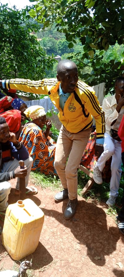 A man in a yellow and black jacket gestures outward, surrounded by other people. Some sit, adorned in colorful clothing and head coverings. One man holds a machete. A yellow container sits on the ground near a goat. Green trees and a distant landscape are visible behind the group, suggesting an outdoor setting. The ground is a reddish-brown soil. The atmosphere appears lively and social.