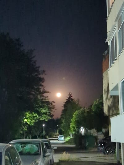 A nighttime scene in Razgrad, Bulgaria, features a bright full moon partially obscured by atmospheric haze, creating a soft glow in the dark sky. To the right, a multi-story building with cream-colored siding and windows is visible. In front of the building, several parked cars are present, including a dark-colored SUV on the right and a silver minivan or station wagon on the left. Trees with illuminated foliage are scattered throughout the scene, casting shadows and adding to the nocturnal ambiance. A streetlamp with a white light is positioned between the trees and the parked cars. In the background, a light-colored vehicle is visible on a road. The overall impression is a quiet residential street at night, illuminated by the moon and artificial lights.