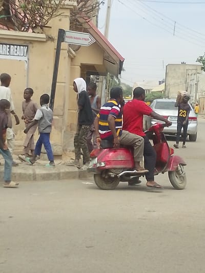 The image captures a detailed urban street scene on Nasiru Kabara Street in Babban Giji, Nigeria, under an overcast sky. In the midground, two adult males are positioned on a red, worn Vespa-style scooter, facing towards the right. The driver wears a red short-sleeved shirt, dark trousers, and brown flip-flops. The passenger, seated directly behind the driver, wears a multi-striped short-sleeved shirt (red, white, blue, yellow), light brown trousers, and brown flip-flops.

To the left of the scooter, on a raised concrete sidewalk bordering a light-colored building, a group of approximately five children and young males are in various stages of movement. A young male in a white hooded sweatshirt and camouflage-patterned trousers is walking from right to left. Adjacent to him, another young male wears a light brown, traditional-style tunic. Further left, a younger boy is visible in a grey vest over a white shirt, blue pants, and light green flip-flops, walking from right to left. On the building's facade, a partial white sign with black text reads "ARE READY". A black pole with a white, arrow-shaped sign points to the right, clearly indicating "NASIRU KABARA STREET".

In the background on the right side of the paved street, a silver sedan is visible. To the right of the sedan, a young male wearing a black t-shirt with the number "23" in yellow and black shorts walks towards the right, carrying a rectangular, light-colored box on his head. Overhead power lines traverse the upper right quadrant of the scene.