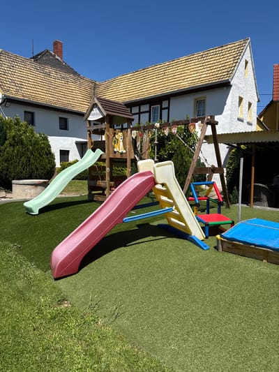 The image shows a backyard playground with a slide, swing set, and a covered sandbox. The playground is situated on artificial turf. In the background is a traditional German building with a tiled roof, half-timbered construction, and white stucco walls. The location is likely Zürchau, Germany. No people are visible in the image.