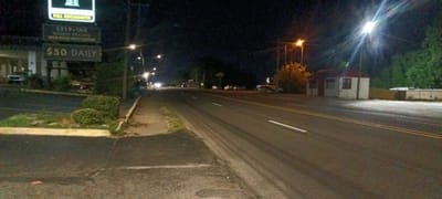The image depicts an urban street scene at night. The location is identifiable by the business sign to the left, which indicates "Full Kitchenette," "$219+TAX Weekly Pricing," and "$50 DAILY." The street is a paved road with streetlights illuminating the area. The road has two lanes on each side, separated by a white line. Visible tire tracks are present in the lanes. On the left side of the road is a sidewalk with foliage and a building further back. On the right side is a small white structure with a red roof, set back from the road. Further down the road, headlights from distant vehicles can be seen. The environment suggests a commercial or residential area in Fort Smith, Arkansas, United States.