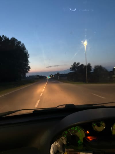 The image captures a view from inside a car on a road at dusk. The road ahead is illuminated by the car's headlights and streetlights, with white dashed lines marking the lanes. To the left, a silhouette of trees forms the edge of the road. In the distance, there are blurred lights, suggesting buildings or other vehicles. The sky is a gradient of deep blue and lighter hues, with a faint crescent moon visible in the upper left corner. The car's dashboard is visible in the foreground, with a speedometer displaying a speed of approximately 100 km/h and a warning triangle illuminated. The scene appears to be in the city of Ponteterra, Italy.