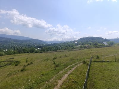This is an outdoor, eye-level, full shot image of a hilly, green landscape on a clear day. The foreground is a grassy meadow with a dirt path winding through it. To the right, a wire fence encloses a section of the meadow. In the midground, there is a line of wooden fence, separating the meadow from a forested area. Scattered among the trees are several houses, some with red roofs and others with darker roofs. The background features rolling hills covered in trees, with a distant body of water visible. The sky is blue with scattered white clouds. The scene is tranquil and suggests a rural setting in or around Nikortsminda, Georgia. There are no people visible in the image.