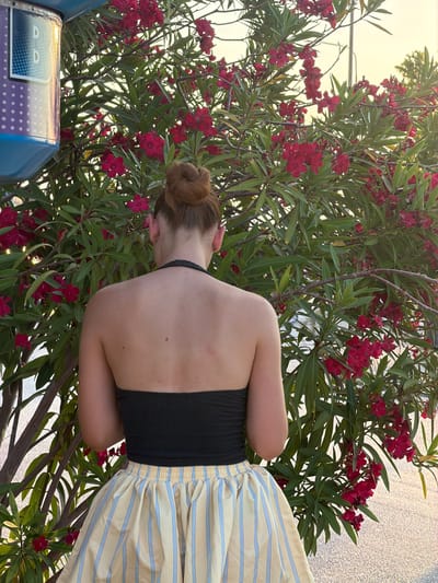 A young woman with her back to the camera is shown from the waist up, with her hair pulled into a bun. She is wearing a black halter top and a yellow skirt with blue stripes. She is standing in front of a flowering tree with red blossoms and green leaves, possibly an oleander. The setting appears to be outdoors in Keramoti, Greece, as indicated by the location context. The lighting suggests it is late afternoon or early evening, with the sun casting a warm glow. The scene is peaceful and serene, with the woman seemingly enjoying the natural surroundings. A portion of a blue object with a patterned design and what appears to be text is visible in the lower left corner of the image.