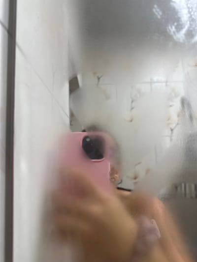 The image is a selfie taken in a bathroom, likely a shower, given the condensation on the mirror. The foreground shows a pink smartphone with a camera module, held by a person whose hand and arm are visible. The phone's case is pink. The person is wearing a light-colored garment, possibly a top, with a textured band visible on the arm, which appears to be a scrunchie. The background is a mirror covered in condensation, with some faint reflections of the environment. The tiling of the shower wall is partially visible on the left side of the image. There are also faint, abstract shapes on the mirror, possibly reflections of decorative elements or a person standing behind the camera. The overall scene is blurry and indistinct due to the condensation. The location in the city of Tryavna, Bulgaria, is not evident from the image itself.
