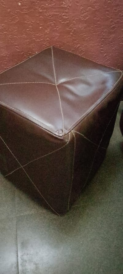 This media file shows a brown ottoman positioned on a tiled floor. The ottoman has a diamond-stitched pattern on its top surface and diagonal stitching on its sides, creating a segmented appearance. The material appears to be faux leather or a similar synthetic fabric, with a slight sheen. The background features a textured, reddish-brown wall, possibly wallpaper or painted concrete with an embossed pattern.
The lighting suggests an indoor setting, likely during the daytime, with the light source creating subtle highlights and shadows on the ottoman. The floor tiles are a neutral, speckled gray-green. There are no people or visible text in the image. The scene is static, with no activity taking place, presenting the ottoman as the sole focus. The context provided indicates the location is Gboko, Nigeria. The overall impression is a simple, functional piece of furniture in a domestic or commercial interior space.