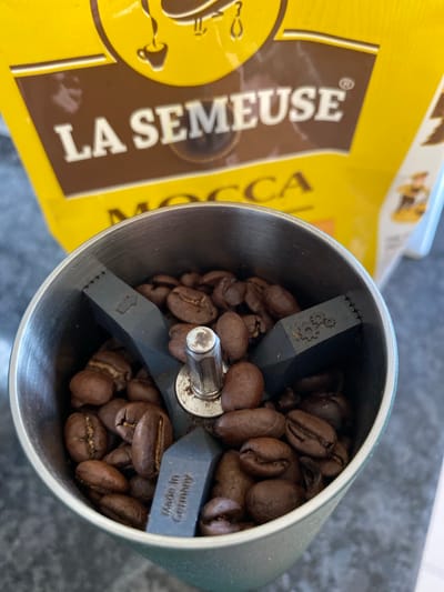 This is a close-up, top-down view of a manual coffee grinder filled with whole coffee beans. The grinder is cylindrical and made of brushed metal. Inside, dark brown coffee beans are visible, some resting on the blades of the grinder. The blades appear to be made of dark grey plastic, with a textured edge and a detail resembling gears etched onto one blade. Another blade has the text "Made in Germany" stamped on it. To the left of the grinder is a partially visible yellow and brown bag, which appears to be a coffee bag. The text "LA SEMEUSE" is prominently displayed in white letters on a brown section of the bag, with a circular logo featuring a stylized human figure. The setting is likely a kitchen countertop, given the dark, textured surface visible in the background. No people are visible, and the scene appears to be lit by natural light, suggesting daytime. The overall impression is of a domestic setting focused on coffee preparation.