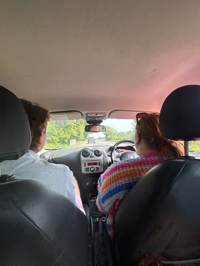 Inside a car, a person with ginger hair and a colorful sweater drives. A passenger sits to their right, leaning back with their head on the headrest. The car is parked on a street with trees and buildings in the background. The car's dashboard and console are visible, showing the radio and other controls. The rearview mirror reflects the driver. The image is taken from the backseat.