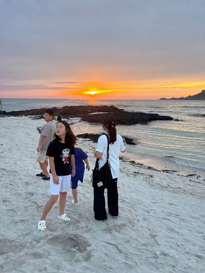 This media file depicts a group of four young people on a sandy beach at sunset. The scene is set in Jeju, South Korea, as indicated by the location context. The sky is ablaze with the warm hues of orange and yellow, reflecting on the gentle waves lapping at the shore. Dark, volcanic rocks punctuate the coastline, contrasting with the soft sand.

Two individuals are standing in the foreground, facing away from the camera. One person, wearing a black t-shirt with a cartoon graphic and white shorts, is looking towards the water, their expression unreadable. The other person, wearing a gray t-shirt and glasses, is also looking out at the sea, holding a smartphone. Behind them, a third person in a white t-shirt and black pants stands with a black bag slung over their shoulder. The fourth person, wearing a blue shirt and white shorts, is partially visible between the two individuals in the foreground.

The group appears to be enjoying a leisurely moment, perhaps reflecting on the day or enjoying the picturesque sunset. The overall mood seems peaceful and serene, amplified by the tranquil setting of the beach at dusk. The sand is imprinted with footprints, suggesting recent activity. No visible text is present in the image.