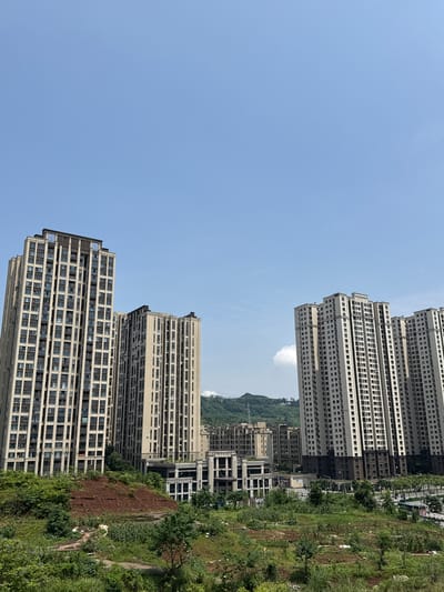 The image presents a scene in Tiaodeng, China, illustrating a blend of dense urban development and natural, hilly landscape under a clear blue sky.
Multiple tall, modern high-rise buildings, likely residential apartment complexes, are prominent. On the left side of the frame, several light-colored towers with numerous, uniformly arranged windows rise dramatically. More similar buildings extend into the mid-ground and background on the right, some appearing newly constructed or still in progress, with sections featuring darker facades or exposed concrete. The architecture is functional and contemporary, typical of growing cities.
The foreground and much of the right side of the image are dominated by a rugged, green landscape. This area features lush grass, various shrubs, and scattered trees, suggesting an uncultivated or recently disturbed natural environment. Patches of reddish-brown soil are visible, indicative of exposed earth, possibly from construction grading or natural erosion on a hillside. Some scattered debris or light trash can be seen within the vegetation. In the far background, distant green hills or mountains are discernible under a bright sky.
The photograph captures a clear, sunny daytime scene, with an expansive, cloudless light blue sky occupying the upper portion of the frame. No people or explicit activities are visible, lending a static quality to the image, which emphasizes the architectural and natural elements. There is no visible text present in the image.