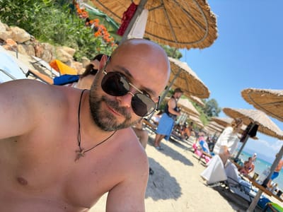 The image shows a man lying on a beach in Greece. He is wearing sunglasses and a small pendant around his neck. In the background, there are several thatched umbrellas providing shade, beach chairs, and other beachgoers. The scene appears to be set on a sunny day, with clear blue skies visible. The sand and the presence of umbrellas and people indicate a typical beach environment. The man's relaxed posture and the overall setting suggest a leisurely day at the seaside.