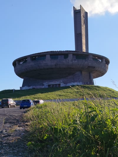 The image shows the Buzludzha Monument, a former communist party headquarters located in the Shipka Pass, Bulgaria. The monument is a large, UFO-shaped structure with a tall tower. It is situated on a grassy hill with a clear blue sky. Several cars are parked on a dirt road in the foreground, and tall grass lines the side of the road. A single person is visible standing on the hill near the monument, looking towards the structure. The monument appears weathered and somewhat dilapidated, with some graffiti visible on its base. The overall scene conveys a sense of abandonment and decay of a once-significant landmark.