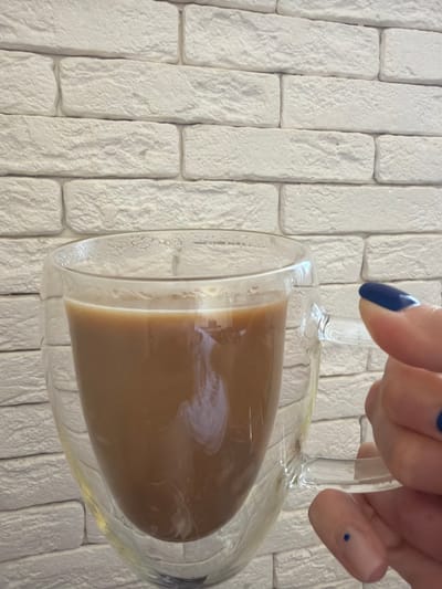 An eye-level medium shot shows a hand with dark blue nail polish holding a clear glass mug filled with a light brown liquid, likely coffee with cream. The mug has a double-walled construction, visible as a slight separation between the inner and outer glass layers. The background is a white brick wall with a textured, rough surface. The lighting is soft and diffused, casting no harsh shadows. The image does not contain any recognizable landmarks or features that would allow for its specific location in Novoxayeyevsk, Russia to be determined.