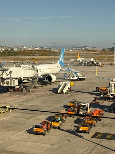 The image captures an airport tarmac in Pendik, Turkey, under a clear blue sky. In the foreground, a white jet bridge extends to connect with a large passenger airplane, which bears the "ajet.com" livery. Various airport service vehicles and baggage carts are spread across the tarmac, some loaded with luggage. In the background, another airplane with yellow tail fins, identified as Pegasus, is partially visible near a jet bridge, with ground crew and service vehicles attending to it. Further in the distance, the cityscape of Pendik is visible, with buildings and urban infrastructure nestled against rolling hills. The scene depicts a typical airport operation with an emphasis on aircraft, ground support equipment, and the urban landscape of Pendik.