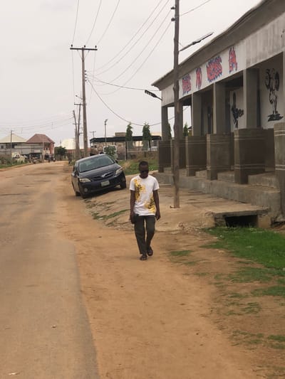 The image shows a street scene in what appears to be a residential or commercial area of Hanwa, Nigeria. A man wearing a white shirt with a gold design and dark trousers is walking along a dirt road or unpaved shoulder of the road. A dark-colored car, possibly a Toyota Corolla, is parked on the side of the road, oriented in the same direction as the man's movement. Overhead power lines stretch across the sky. In the background are buildings, some with corrugated roofing, and what appears to be a partially constructed structure with exposed concrete. The ground is mostly dirt and some patches of grass are visible. The sky is overcast. The image suggests a day time setting.