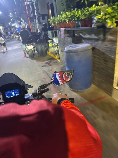 This is a first-person perspective shot from a motorcycle in Manila, Philippines, at night. The rider, wearing a red hooded jacket, is looking forward, with the handlebars and instrument cluster of the motorcycle visible. The instrument cluster displays a time of 8:11 and a speed of 9 km/h. A decorative rearview mirror with a pattern resembling a flag is attached to the handlebars.

The scene is a street, likely in a busy urban area, given the presence of various objects and structures. To the left, a large blue barrel sits near a wooden structure with some greenery. Further in the background, a glimpse of other motorcycles and people can be seen, suggesting a bustling environment. The lighting indicates it's nighttime, possibly with artificial streetlights illuminating the scene. The overall impression is that the motorcycle is stopped or moving slowly in a vibrant, possibly crowded, street. No specific weather conditions are discernible.