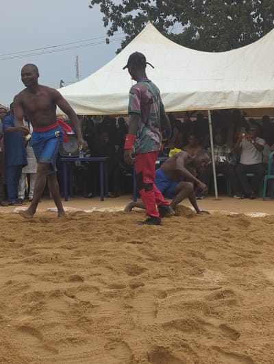 The image shows a wrestling match taking place on a sand-covered surface in Kafanchan A, Nigeria. Two shirtless men wearing shorts, one blue and one blue with a red belt, are interacting. The man in blue is holding a small microphone while the other is standing next to him wearing a baseball cap. Behind them, a large crowd is seated or standing, partially under a white tent structure. Several people in the crowd are using mobile phones, possibly to take pictures or videos. A tree is visible to the left of the tent.