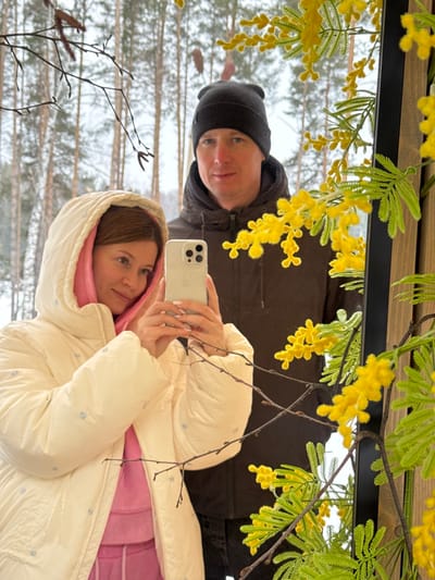 A woman and a man are captured in a mirror selfie within what appears to be an indoor or semi-enclosed space, located in Mishkino, Russia, against a backdrop of a snowy winter forest. The woman, positioned in the foreground and to the left, is Caucasian with light brown hair and wears a cream-colored hooded puffer jacket over a pink hooded sweatshirt. She holds a light-colored smartphone with her left hand, pointed towards the mirror to take the photograph, displaying light pink nail polish. She has a faint smile and gazes towards the phone. The man, a Caucasian individual with short light brown hair, stands behind and slightly to the right of the woman. He wears a black beanie with a small white logo and a dark brown or black winter jacket, maintaining a neutral expression while looking towards the reflected camera. The mirror itself has a thin black frame visible on the right. Decorative bright yellow mimosa-like flowers with small green leaves are arranged prominently in the foreground and on the right side, partially obscuring the man and the reflected environment. Several bare, dark branches are also visible in the immediate foreground. The reflection in the mirror reveals a dense winter forest environment characterized by tall conifer trees, a few bare deciduous trees, and a ground uniformly covered in snow under an overcast sky.