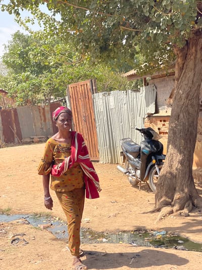 An adult female is depicted walking right-to-left on a dirt path in Chikaji, Nigeria, under daylight conditions. She wears a yellow-brown patterned top and matching trousers, a red and white patterned head covering, and a red and white striped fabric draped over her left shoulder and arm. A white bracelet is visible on her left wrist. She looks directly towards the viewer with a neutral expression. Her left foot is forward, wearing a light-colored sandal.
To her right, a dark motorcycle is parked next to a tree with a thick trunk and exposed roots. Behind the motorcycle and the woman are structures made of corrugated metal sheets, some rusted brown, forming walls or fences. A vertical wooden gate, also brown, is visible behind the woman. Further back, green foliage from trees and bushes is present. A small structure with a light-colored corrugated roof is visible behind the tree and motorcycle. The ground is dry dirt, with several shallow puddles containing dark liquid and scattered debris visible in the foreground. Shadows are cast on the ground from the tree and other unseen elements, indicating strong overhead sunlight.