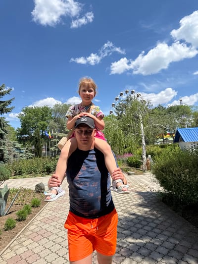 A man and a child are on a paved pathway with an ornate pattern. The man is carrying the child on his shoulders. The man wears a dark cap, a dark t-shirt with a design, and bright orange shorts. The child wears a printed shirt and pink shorts. The man looks at the camera and smiles. The child looks at the camera. In the background, there is a large Ferris wheel, green trees, and a blue roof. Above, there is a blue sky with white clouds. This photo was most likely taken in Yevpatoriya, Ukraine.