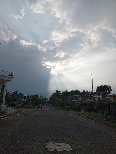 This image depicts a residential street scene in the city of Weintiapa, Liberia, observed under a partially cloudy sky with visible sunbeams. The upper two-thirds of the frame are occupied by a sky exhibiting dark grey storm clouds on the left transitioning to brighter clouds on the right, from which crepuscular rays emanate downward, indicating the sun breaking through. Atmospheric moisture or light rain streaks are visible in the upper left quadrant.

The lower third of the image shows a wide, dark-paved road constructed of interlocking pavers or cobblestones, receding into the distance. A light-colored, rectangular patch is present on the road surface in the immediate foreground.

On the left side of the road, the corner of a multi-story white building with classical columns and a dark, ornate metal balcony railing is prominently featured. Further down, several smaller buildings with grey roofs are partially obscured by trees.

The right side of the road is lined with residential buildings, lush green trees, and shrubs. A yellow building with a red-tiled roof is visible on the mid-right. A black metal fence runs along the right-hand properties, separating them from the street. A modern street lamp with a curved arm stands on the right side of the road in the mid-ground.

In the middle distance on the road, a dark-colored SUV is visible, facing away from the viewer. Further beyond, smaller vehicles, possibly motorbikes, are faintly discernible. Dense green foliage in the background suggests hills or mountains. No individuals are clearly visible.