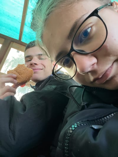 Two individuals are positioned closely together in an upside-down selfie taken at an upward angle. The individual on the left is wearing glasses and has fair hair. The individual on the right is holding a partially eaten sandwich. The background is comprised of an outdoor scene with some vegetation. The location is Karavelovo, Bulgaria.