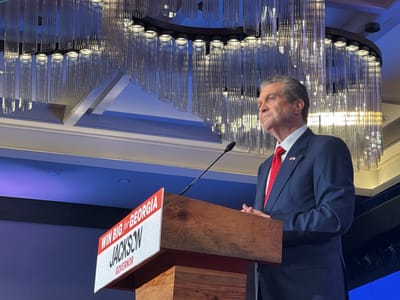 The image shows a man in a suit standing behind a podium. The podium has a sign that says "WIN BIG for GEORGIA JACKSON GOVERNOR". The man is wearing a red tie and a dark blue suit. The background is a blue wall and a large, modern chandelier. The scene appears to be taking place indoors, potentially in a ballroom or event space in Atlanta, United States. The man is speaking at the podium.