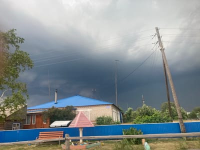 The image depicts a rural scene in Kormikha, Russia, under a dramatically overcast sky. A dark, foreboding storm cloud looms overhead, casting a somber mood. In the foreground, a bright blue fence runs horizontally across the frame, creating a distinct visual separation. To the left of the fence, a wooden bench with orange slats is positioned on a patch of grass. Behind the fence, several structures are visible. A building with a blue metal roof and a brick facade stands prominently, with trees and foliage surrounding it. To the left of this building, a portion of a wooden house with a visible window is present. Various utility poles and wires are scattered throughout the scene, with one particularly tall wooden pole dominating the right side of the frame. A small, pink, pyramid-shaped structure, possibly a child's playhouse or decorative element, is situated near the blue fence. The overall impression is one of an impending storm in a quiet, rural settlement. There are no people or animals visible in the image.
